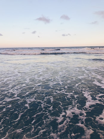 A serene ocean scene with gentle waves rolling towards the shore under a clear sky. The horizon separates the sea from a pale sky scattered with a few clouds, creating a tranquil and peaceful atmosphere.