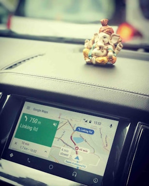 A small Ganesha idol is placed on the dashboard of a car, next to a screen displaying Google Maps navigation. The map shows a route with distance and time details.