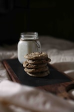 A stack of homemade chocolate chip cookies is arranged on a wooden board next to a glass jar filled with milk. The background is dimly lit, creating a cozy and inviting atmosphere.