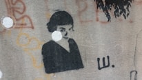 A graffiti-style stencil art on a textured wall depicts a monochromatic portrait of a person with dark hair against a light background. Surrounding the portrait are abstract shapes and patterns in various colors such as orange and red.