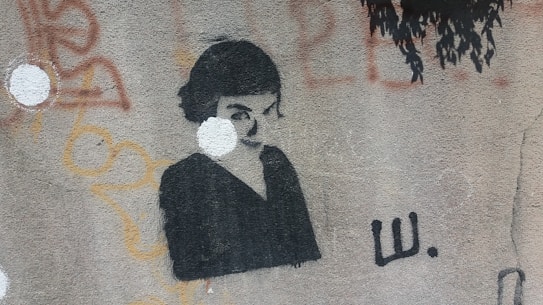 A graffiti-style stencil art on a textured wall depicts a monochromatic portrait of a person with dark hair against a light background. Surrounding the portrait are abstract shapes and patterns in various colors such as orange and red.