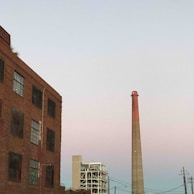 An industrial scene with a tall chimney rising into a pastel-colored sky, adjacent to a large, brick building with several windows. Another industrial structure with a complex scaffolding is visible in the distance. Electrical lines cut across the sky, adding to the urban, industrial atmosphere.