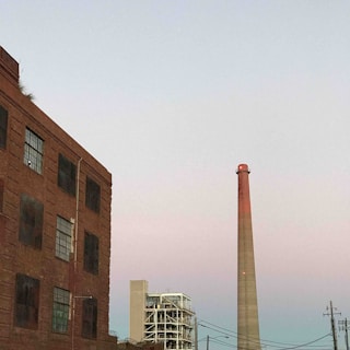 An industrial scene with a tall chimney rising into a pastel-colored sky, adjacent to a large, brick building with several windows. Another industrial structure with a complex scaffolding is visible in the distance. Electrical lines cut across the sky, adding to the urban, industrial atmosphere.