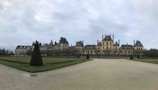 A large, symmetrical chateau with a classic architectural style, featuring numerous chimneys and dormer windows, surrounded by a neatly landscaped garden with trimmed hedges and a wide path leading up to the building. The sky is overcast, creating a subdued and tranquil atmosphere.