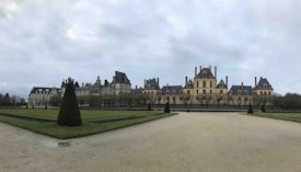 A large, symmetrical chateau with a classic architectural style, featuring numerous chimneys and dormer windows, surrounded by a neatly landscaped garden with trimmed hedges and a wide path leading up to the building. The sky is overcast, creating a subdued and tranquil atmosphere.