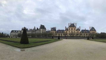 A large, symmetrical chateau with a classic architectural style, featuring numerous chimneys and dormer windows, surrounded by a neatly landscaped garden with trimmed hedges and a wide path leading up to the building. The sky is overcast, creating a subdued and tranquil atmosphere.