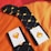 A pair of black socks with a geometric diamond pattern in yellow and orange, laid out on an orange and red surface. The socks have white cards attached displaying an orange diamond logo and a website URL, providing a contrasting and vibrant look.
