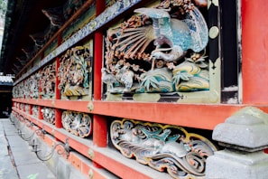 Intricate and colorful carvings adorn a traditional Japanese building, featuring detailed depictions of animals and nature motifs. The designs are crafted into the wooden structure, highlighted by vibrant shades and are framed by bold red painted beams.