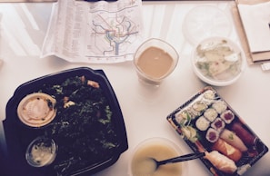 A variety of food items including a green salad in a black container with dressing, a clear cup with a beverage, a bowl of soup with a spoon, a container of mixed sushi rolls, and a small salad in a separate bowl are arranged on a table. A map with subway routes is laying open in the background.