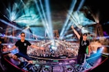 A vibrant concert scene with two DJs on stage, surrounded by a massive, energetic crowd. The stage is illuminated with colorful lights and impressive visual effects, creating a dramatic atmosphere. Laser beams and mist add to the dynamic setting.