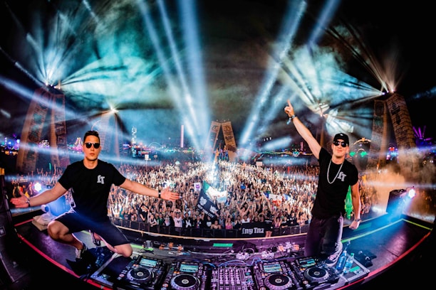 A vibrant concert scene with two DJs on stage, surrounded by a massive, energetic crowd. The stage is illuminated with colorful lights and impressive visual effects, creating a dramatic atmosphere. Laser beams and mist add to the dynamic setting.