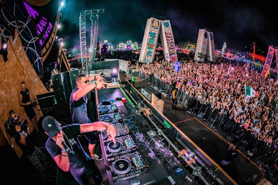 A large crowd of people is gathered at a music festival. Two DJs are performing on a stage packed with audio equipment. The vibrant scene includes colorful lights in the background and various flags being waved by the audience.