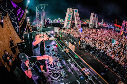 A large crowd of people is gathered at a music festival. Two DJs are performing on a stage packed with audio equipment. The vibrant scene includes colorful lights in the background and various flags being waved by the audience.