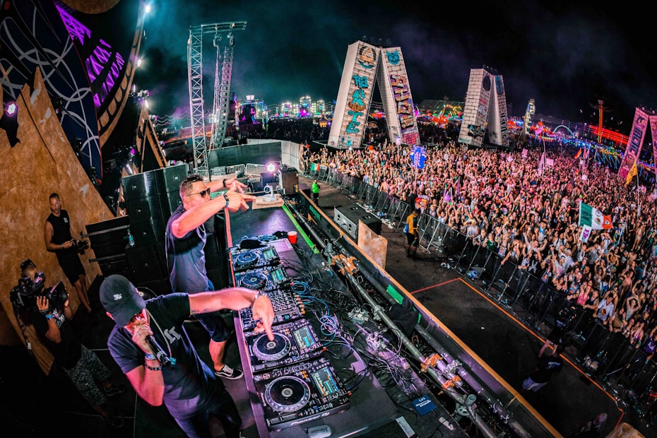 A large crowd of people is gathered at a music festival. Two DJs are performing on a stage packed with audio equipment. The vibrant scene includes colorful lights in the background and various flags being waved by the audience.