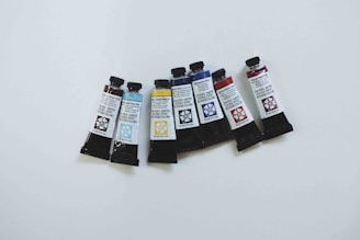 Seven tubes of Daniel Smith Extra Fine Watercolors are arranged in a slight curve on a white surface. The tubes feature various colors and corresponding colors printed on the labels, including shades of brown, blue, yellow, and red. Each tube has a dark cap and a floral design next to their labels.