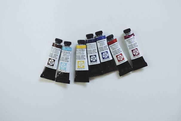 Seven tubes of Daniel Smith Extra Fine Watercolors are arranged in a slight curve on a white surface. The tubes feature various colors and corresponding colors printed on the labels, including shades of brown, blue, yellow, and red. Each tube has a dark cap and a floral design next to their labels.