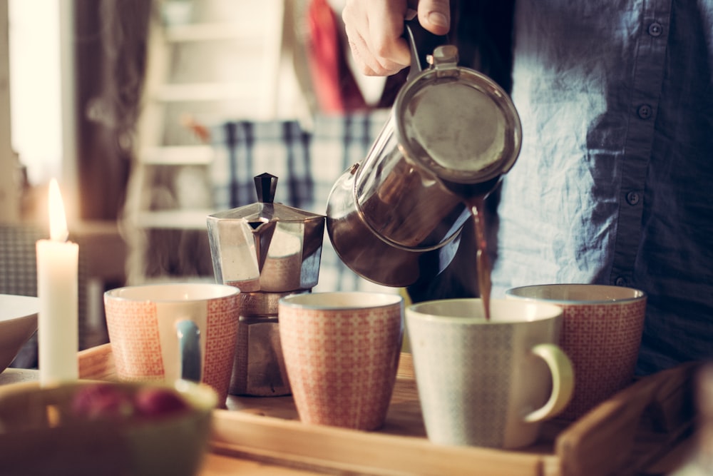 person pouring tea on white ceramic mug using teapot beside moka pot