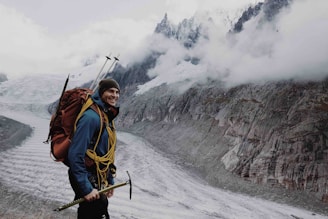A person is standing on a rocky mountainous terrain, dressed in mountain climbing gear. The individual has a large backpack, climbing tools, and ropes. In the background, there are snowy peaks partially covered by clouds and a glacier running through the valley.