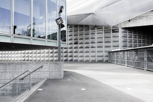 A modern architectural setting with large glass and metal structures. The scene includes geometric facades with reflective surfaces, an outdoor area with concrete and asphalt, and metal fences. A lone light pole and stairs descending into an underground space add to the urban landscape.