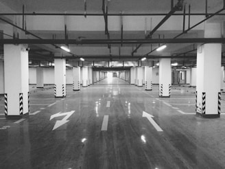 A spacious and empty underground parking lot, with numerous parking spaces and pillars supporting the ceiling. Striped patterns are visible on the pillars, while directional arrows are painted on the smooth, reflective concrete floor. Overhead lighting illuminates the area brightly, creating a sense of openness.