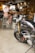 A classic-style motorcycle with a shiny black and chrome finish is parked indoors. In the background, a person wearing an off-the-shoulder top and light brown pants is seated at a high table, suggesting a casual cafe setting. The motorcycle features intricate details in its design and appears well-maintained.