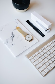 A workspace setup featuring a marble-patterned notebook, a gold wristwatch with a white face, a black and white ceramic cup, a white stapler, a black ruler with numbered markings, and a slim, white and gold keyboard.