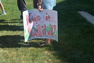 A person is holding a handmade sign with colorful text saying 'Keep it Wild!' on a grassy area. The sign includes drawings of trees and zigzag patterns in various colors.