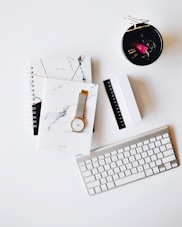 Apple Magic Keyboard near round gold-colored analog watch with silver-colored mesh band with notebook on white surface photography