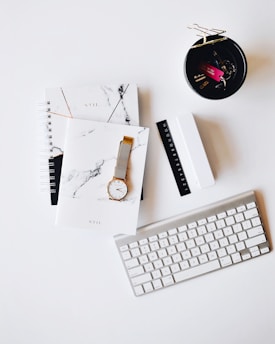 Apple Magic Keyboard near round gold-colored analog watch with silver-colored mesh band with notebook on white surface photography