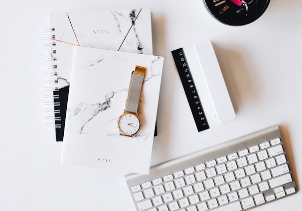 Apple Magic Keyboard near round gold-colored analog watch with silver-colored mesh band with notebook on white surface photography