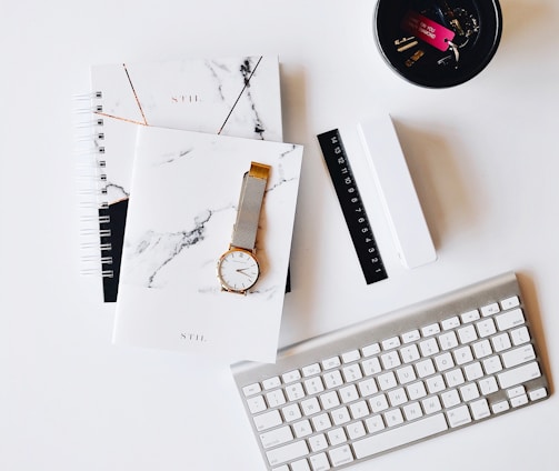 Apple Magic Keyboard near round gold-colored analog watch with silver-colored mesh band with notebook on white surface photography