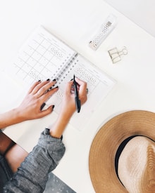 An overhead view of a person writing in a planner or calendar on a white desk. Other objects on the desk include a wide-brimmed hat, two metallic binder clips, and a clear case with a roll of washi tape.