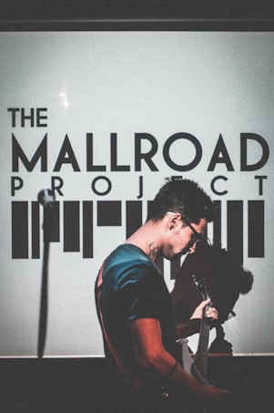 A person is playing a guitar in front of a backdrop with text that reads 'The Mallroad Project'. The scene is dimly lit, casting a dramatic shadow behind the musician.