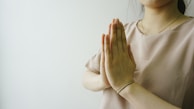 Hands are positioned in a prayer gesture near the chest, with a soft focus on the upper body. The person is wearing a light beige blouse and a simple bracelet on the wrist. The background is a light, minimalist setting, adding a serene feel to the composition.