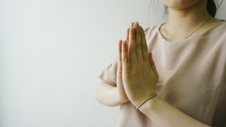 Hands are positioned in a prayer gesture near the chest, with a soft focus on the upper body. The person is wearing a light beige blouse and a simple bracelet on the wrist. The background is a light, minimalist setting, adding a serene feel to the composition.