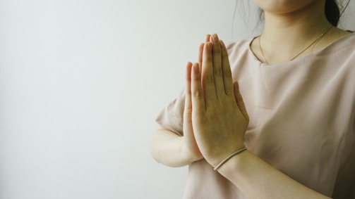 Hands are positioned in a prayer gesture near the chest, with a soft focus on the upper body. The person is wearing a light beige blouse and a simple bracelet on the wrist. The background is a light, minimalist setting, adding a serene feel to the composition.