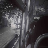 A view from inside a bus looking out the window, showing the street outside with a person walking on the sidewalk and a parked tuk-tuk. The bus interior is dimly lit, with several passengers visible, and the window is open.