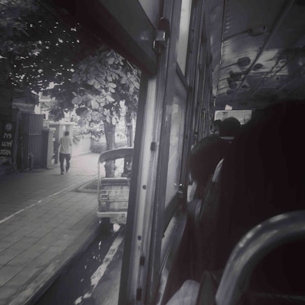 A view from inside a bus looking out the window, showing the street outside with a person walking on the sidewalk and a parked tuk-tuk. The bus interior is dimly lit, with several passengers visible, and the window is open.