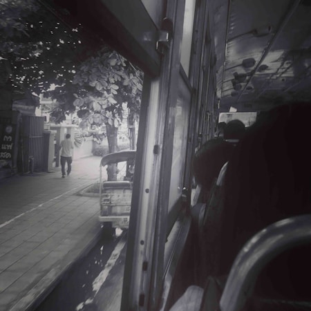 A view from inside a bus looking out the window, showing the street outside with a person walking on the sidewalk and a parked tuk-tuk. The bus interior is dimly lit, with several passengers visible, and the window is open.