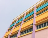 A multicolored building facade with several floors, featuring vibrant hues of yellow, teal, pink, and purple. Each floor is sectioned, with long rectangular windows lining the walls, surrounded by colorful panels.