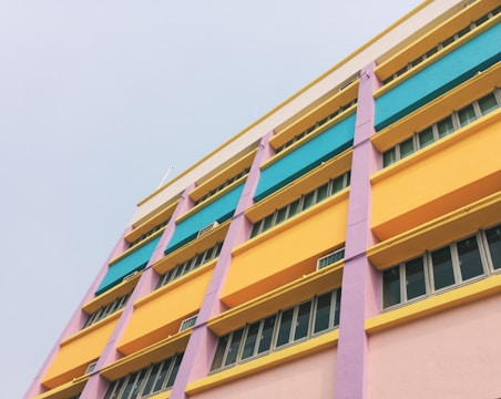 A multicolored building facade with several floors, featuring vibrant hues of yellow, teal, pink, and purple. Each floor is sectioned, with long rectangular windows lining the walls, surrounded by colorful panels.
