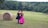 Two people, dressed in contrasting vibrant dresses, walk arm in arm through a lush green field dotted with large hay bales. The field is bordered by dense forest with various shades of green trees. One person wears a bright pink gown, while the other wears a black outfit. The scene conveys a feeling of serenity and companionship.