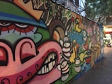 A colorful, urban graffiti mural featuring abstract faces and various cartoonish characters with exaggerated expressions. The artwork spans a long section of a city wall, blending vibrant colors and playful imagery. The surrounding area includes trees and buildings, which indicate an urban environment.