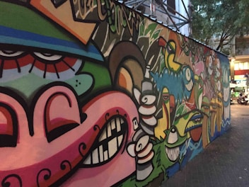 A colorful, urban graffiti mural featuring abstract faces and various cartoonish characters with exaggerated expressions. The artwork spans a long section of a city wall, blending vibrant colors and playful imagery. The surrounding area includes trees and buildings, which indicate an urban environment.