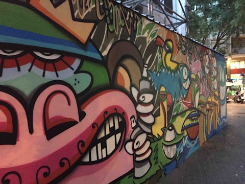 A colorful, urban graffiti mural featuring abstract faces and various cartoonish characters with exaggerated expressions. The artwork spans a long section of a city wall, blending vibrant colors and playful imagery. The surrounding area includes trees and buildings, which indicate an urban environment.
