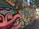 A colorful, urban graffiti mural featuring abstract faces and various cartoonish characters with exaggerated expressions. The artwork spans a long section of a city wall, blending vibrant colors and playful imagery. The surrounding area includes trees and buildings, which indicate an urban environment.