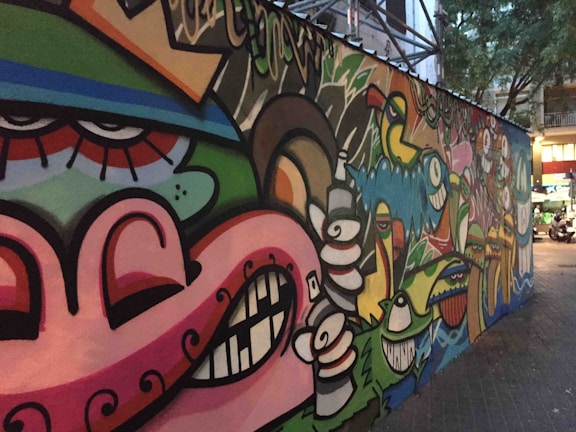 A colorful, urban graffiti mural featuring abstract faces and various cartoonish characters with exaggerated expressions. The artwork spans a long section of a city wall, blending vibrant colors and playful imagery. The surrounding area includes trees and buildings, which indicate an urban environment.