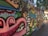 A colorful, urban graffiti mural featuring abstract faces and various cartoonish characters with exaggerated expressions. The artwork spans a long section of a city wall, blending vibrant colors and playful imagery. The surrounding area includes trees and buildings, which indicate an urban environment.