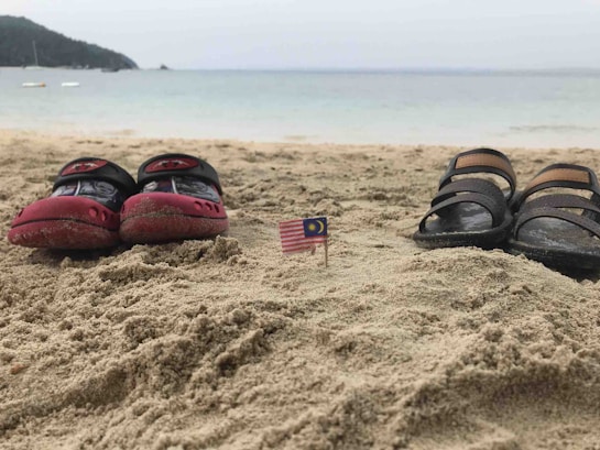A sandy beach with two pairs of shoes placed facing the sea; a small Malaysian flag is stuck in the sand between the shoes. The background shows a calm sea with a slightly overcast sky and distant landforms.