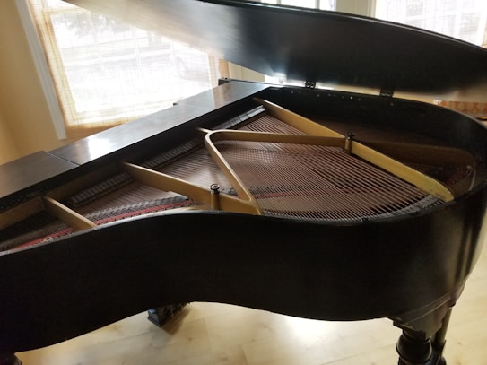 A grand piano with its lid partially open revealing the strings and hammers inside. Natural light filters through a window with blinds, casting soft shadows onto the piano and the wooden floor.